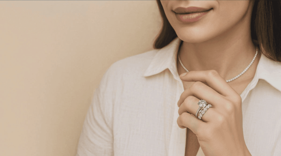 a woman against a beige wall wearing a neutral button down blouse wearing a diamond tennis necklace and showing her engagement ring and matching wedding ring